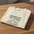 Folded t-shirt with 'Raised by the Islands' text on a wooden surface