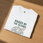 White t-shirt with 'Raised by the Islands' text and graphics on a wooden surface